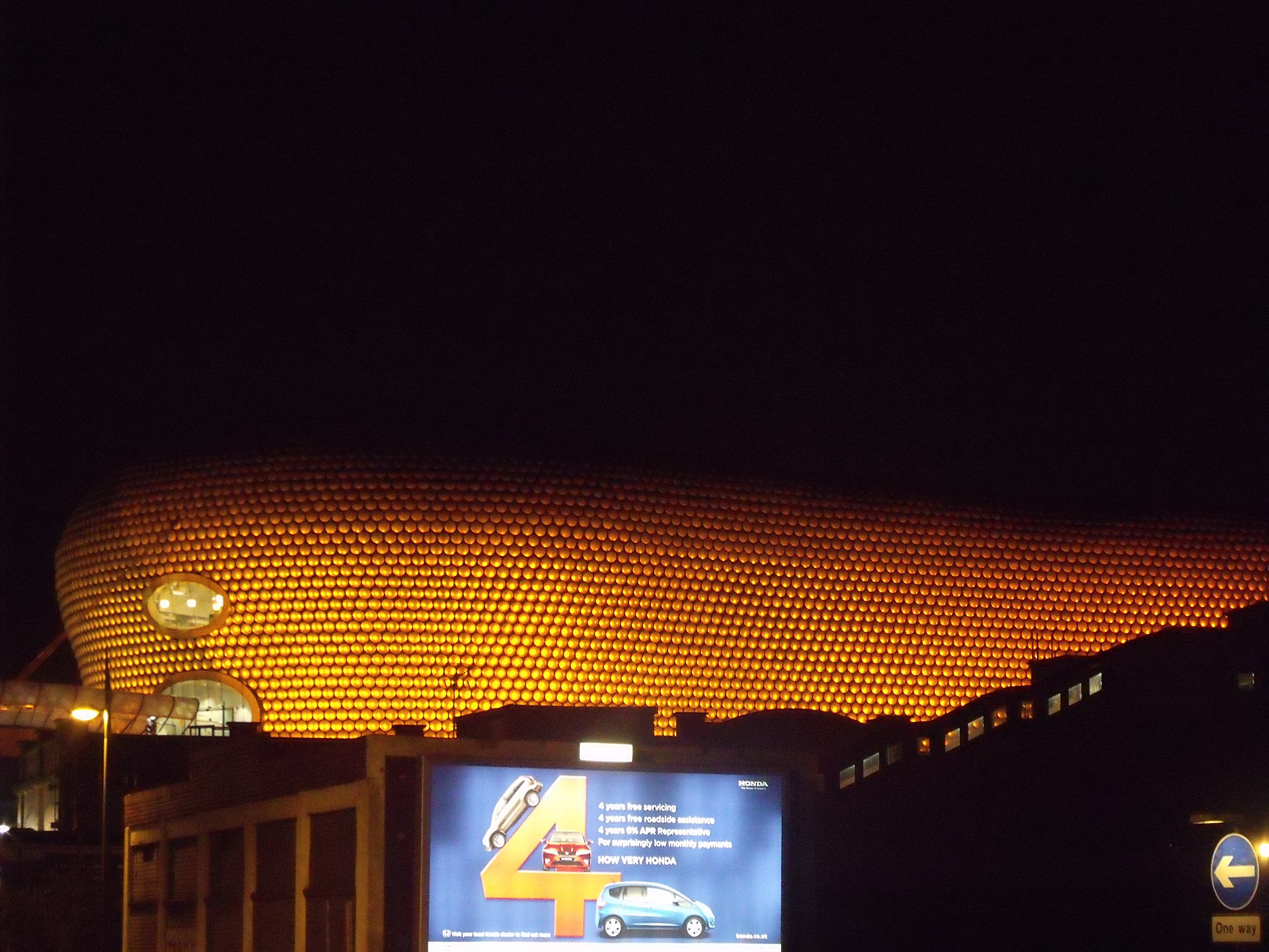 Painel publicitário iluminado em rua à noite, com fachada dourada ao fundo