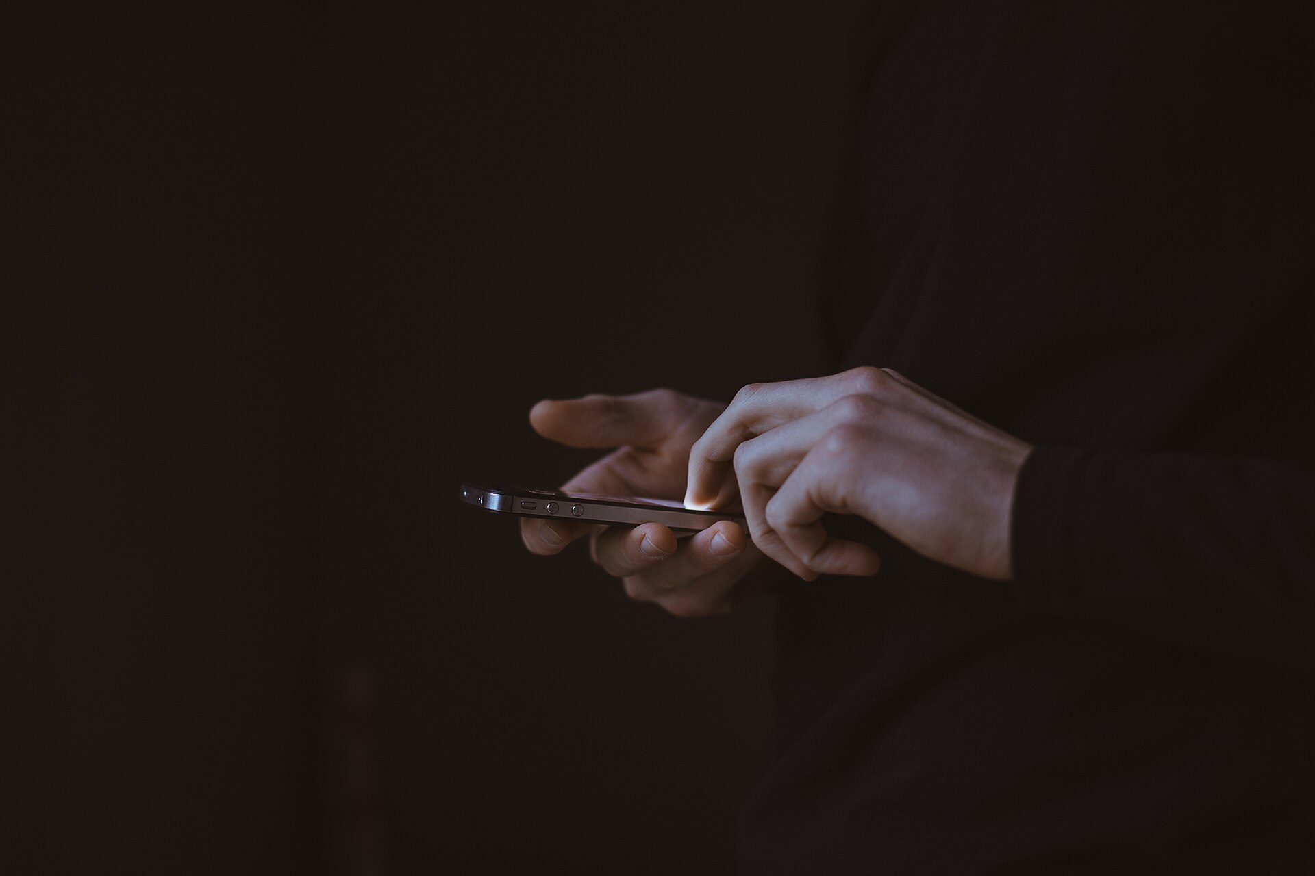 Mãos digitando em um celular, iluminadas pela tela em um ambiente escuro